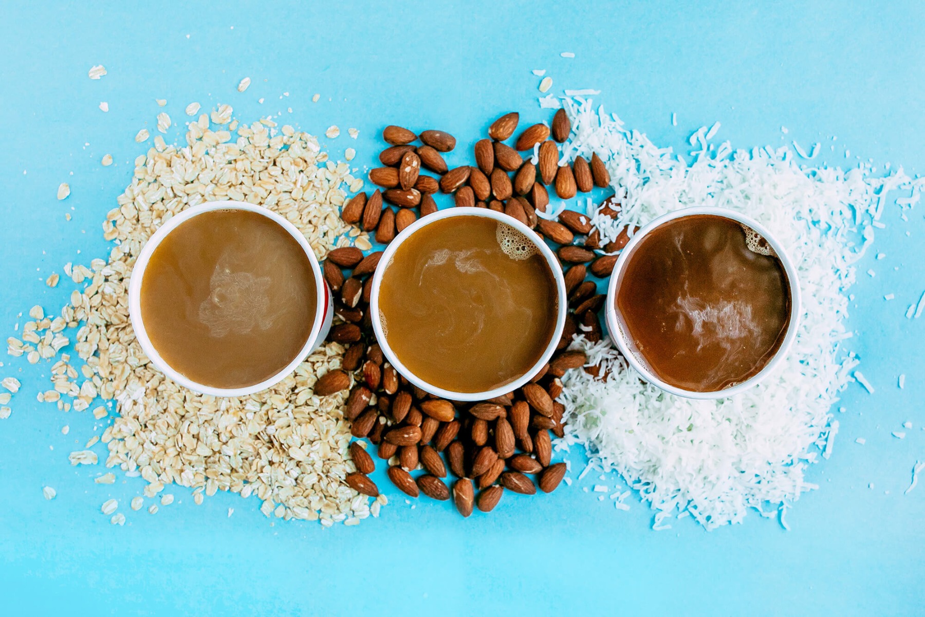 Top shot of three cups of coffee sitting on blue table with oats, almonds and coconut